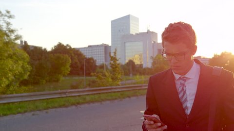 CLOSE UP, SUPER SLOW MOTION, SUN FLARE: Businessman reading a text and getting bad news while walking home at sunset. Caucasian yuppie is surprised and worried as he reads a text on his way from work.