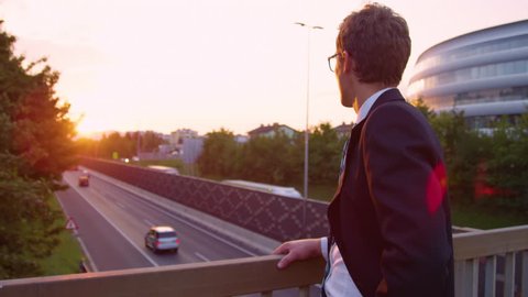 SLOW MOTION, LENS FLARE, CLOSE UP: Young Caucasian businessman lost in thought watches the busy highway below him at sunrise. Yuppie thinking about his stressful job while observing the golden sunset.