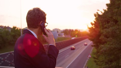 SLOW MOTION, LENS FLARE, CLOSE UP: Young businessman talking on his phone and watching the cars speed down the highway below him. Unrecognizable yuppie calling girlfriend he will be coming home late.