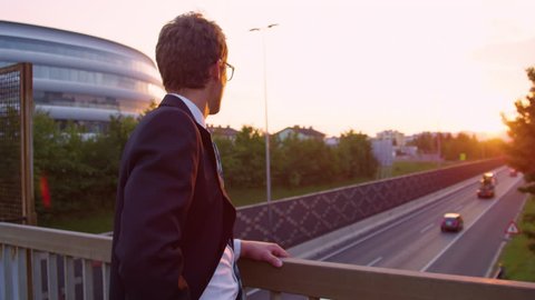 LENS FLARE, CLOSE UP, SLOW MOTION: Unrecognizable manager thinking and watching cars driving down freeway below him. Young businessman lost in thought as he watches the sunset illuminating the city.