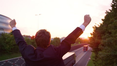 CLOSE UP, SLOW MOTION, SUN FLARE: Golden morning sunbeams shine on excited businessman standing above the highway with outstretched arms. Unrecognizable yuppie celebrating a successful work meeting.