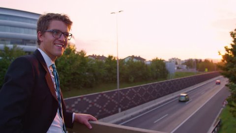 SLOW MOTION SUN FLARE PORTRAIT: Cheerful yuppie looking into the camera and looks at the sunset above the busy highway running past the urban city. Cinematic shot of businessman observing the freeway.