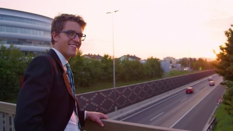 PORTRAIT, SUN FLARE, SLOW MOTION: Successful Caucasian yuppie turns around while watching the sunset above the highway. Young businessman smiles while standing above the busy freeway running past city
