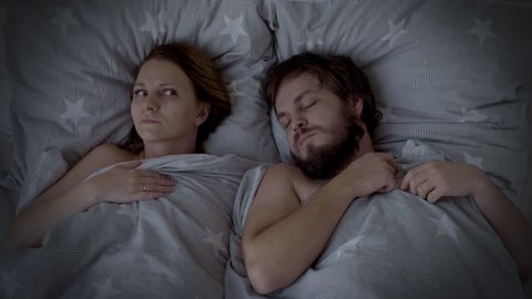 Man Snoring in Bed, Woman cannot sleep. Husband snoring, his Wife is Waking up and closing her ears. Spouses are Sleeping in bedroom at Night.