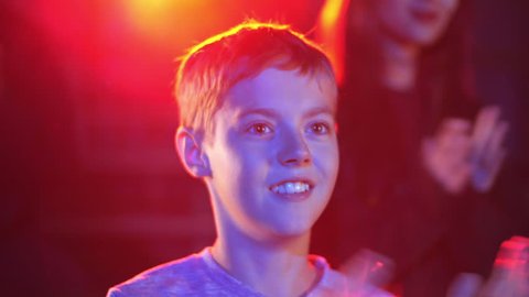 Cheerful caucasian kid clap hands with impressions and smile in spotlights. Successful circus winter tour premiere. Music festival concert for children at holiday time. People applauds at background
