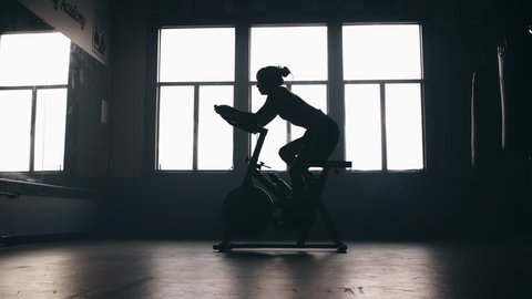 Panning shot of a silhouetted female athlete pedaling on a spin bike in a boxing gym