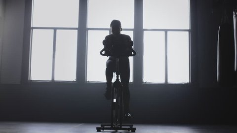 Wide panning shot of a silhouetted woman confidently exercising on a spin bike in a gym