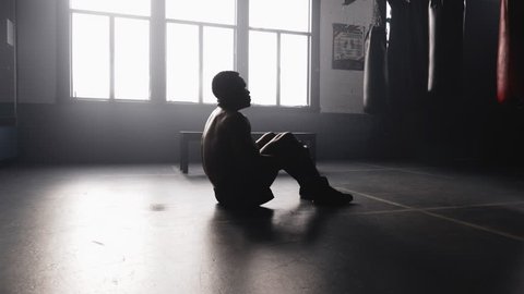 A muscular African American athlete sits on the floor in a boxing gym and takes deep breaths after a work out