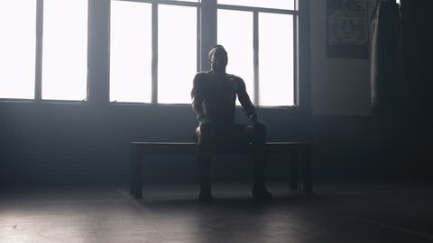 Portrait of a muscular African American athlete sitting on a bench, taking deep breaths in front of a sunlit window in a hazy boxing gym