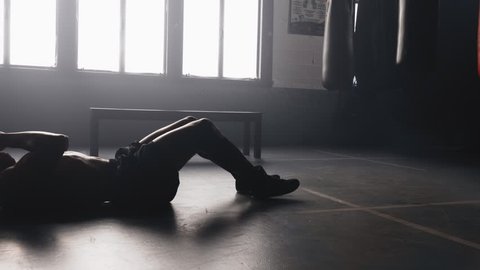 A shirtless African American athlete warms up with sit up punches in a hazy boxing gym. He stops and takes deep breaths