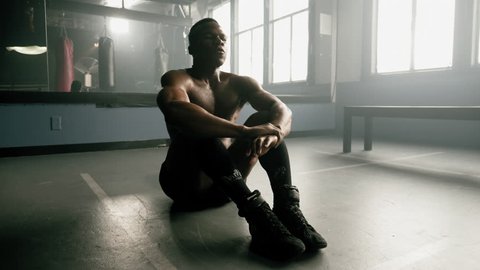 Panning shot of a muscular African American athlete taking a deep breath and then warming up with sit up punches in a boxing gym