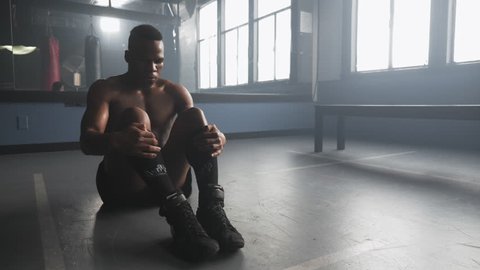 Panning portrait of a sweaty African American athlete sitting on the floor in a boxing gym, and gazing out a sunlit window