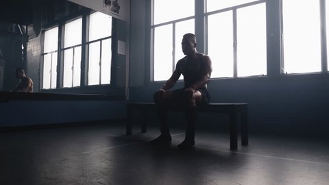 Panning shot of a handsome African American athlete sitting on a bench in an old boxing gym and stretching, reflected in a mirror