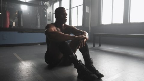 A muscular African American athlete sitting in a boxing gym takes deep breaths after warming up, and looks intensely into the camera