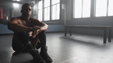 Panning shot of a muscular African American athlete sitting in a boxing gym takes deep breaths after warming up, and looks intensely into the camera