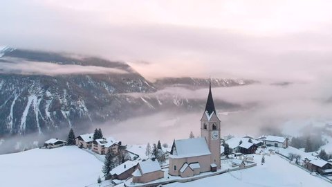Winter top view of the Swiss village with a church, fog and clouds in the mountains in the Alps, snow-covered houses in the valley of mountains and lakes, flying above the clouds and sunset. 