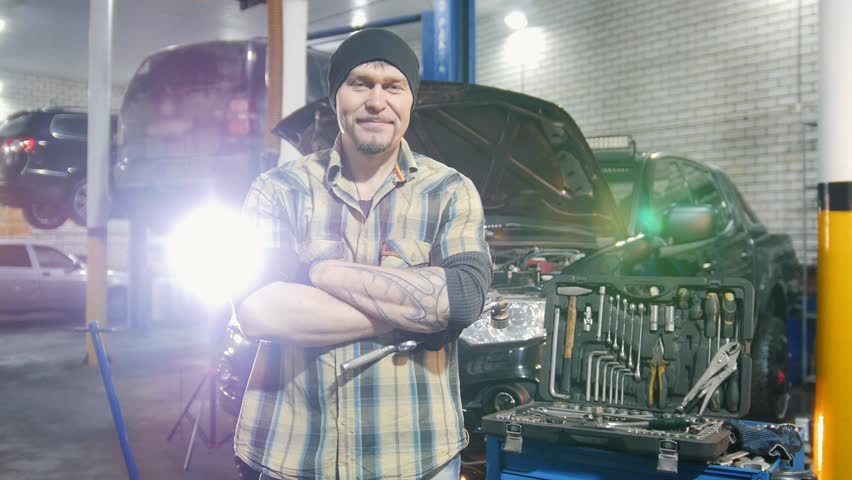 Car service. Brutal mechanic man standing by the car and smiles. Removes the working attributes