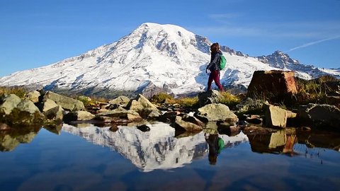 Woman hiker crosses creek with reflection of Mount Rainier in the background. This beautiful peak is a volcano and is in a national park in America.