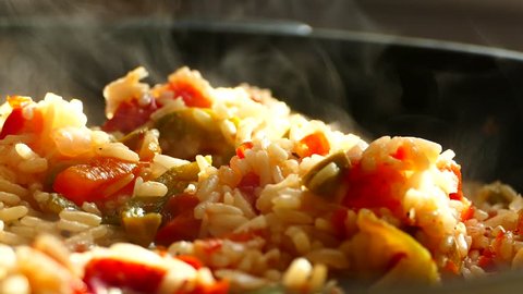 Prepare pilaf in a large frying pan. A dish of rice and pieces of vegetables. Steam comes from a hot lunch. Home kitchen. Pilaf is stirred with a wooden spatula.