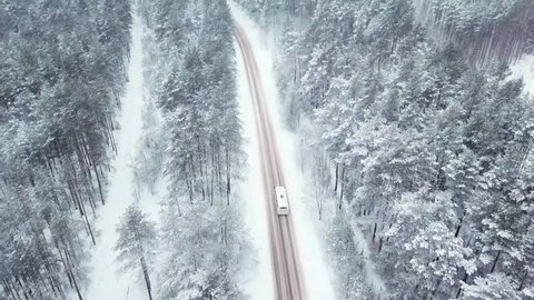 A bus is driving along a snowy road in the middle of a winter frozen forest. Firs and pines in the snow. Snowing. Aerial view
