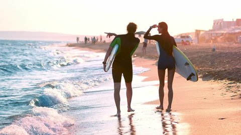 A young couple of guy and girl surfers are walking along the beach in wetsuits. In the hands of holding surfboards. Look at the waves and the sunset.