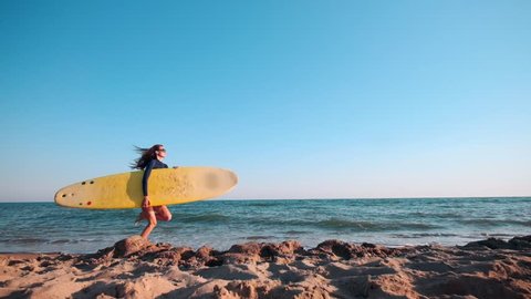 SLOW MOTION: Beautiful slim girl walking on the beach, holding surfboard. Runs in the evening during sunset. Wears a swimming suit wetsuit, protective waterproof suit