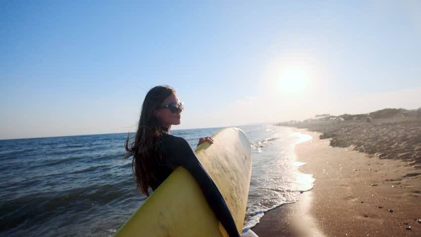 SLOW MOTION: Beautiful slim girl walking on the beach, holding surfboard. Runs in the evening during sunset. Wears a swimming suit wetsuit, protective waterproof suit | Shutterstock HD Video #1022964181