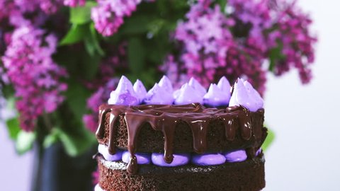 Decorating top of the cake with cream cones, liquid chocolate coating and purple macaroon cakes in front of the lilacs bouquet.   Adlı Stok Video