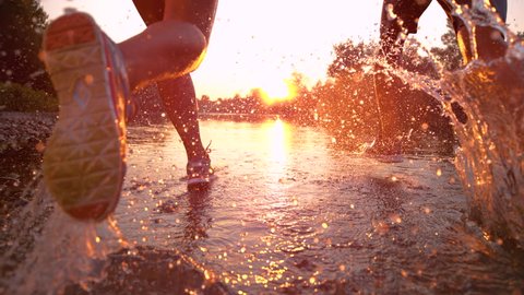 SLOW MOTION TIME WARP, SUN FLARE, CLOSE UP: Fit training partners run in refreshing river water towards beautiful summer sunset. Glassy drops of water fly as young couple jogs through serene nature.