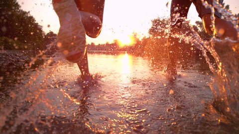 SUPER SLOW MOTION, CLOSE UP, LENS FLARE: Unrecognizable active woman and man running towards the sunset and splashing river water. Carefree sporty couple jogging in the serene nature at golden hour.