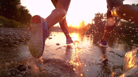 SUPER SLOW MOTION, LENS FLARE, CLOSE UP: Golden sunbeams shine on the fit couple jogging in the tranquil stream. Young woman and man running towards the sunset and splashing the refreshing water.