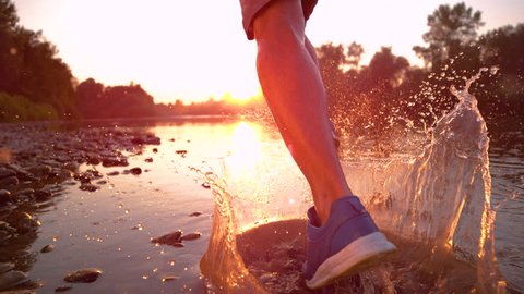 SUPER SLOW MOTION, CLOSE UP, LENS FLARE: Athletic man runs in the shallow stream water towards the sunset. Cinematic shot of golden morning sunbeams shining on sportsman jogging in beautiful nature.