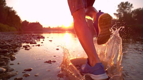 SLOW MOTION TIME REMAP CLOSE UP: Unrecognizable sportsman running in the river shallows at golden hour. Cinematic shot of male jogger splashing stream water as he runs towards the picturesque sunset.