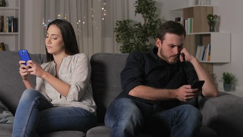 Angry couple using their smartphones after argument sitting on a couch at home in the night