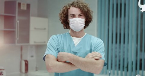 Closeup young doctor or surgeons in a modern clinic room taking off the surgical mask and smiling large in front of the camera , curly hair and white teeth. 4k