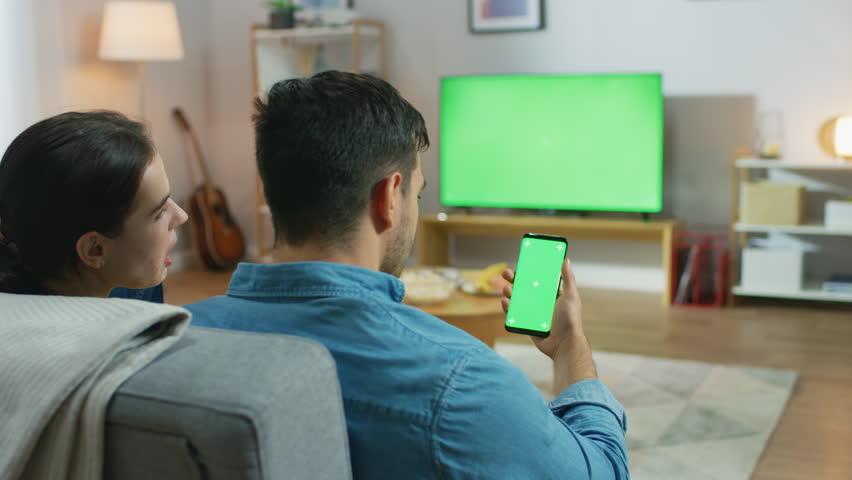 Happy Couple Sitting At Home in the Living Room Watching Green Chroma Key Screen Television, Relaxing on a Couch. Guy also Uses Green Mock-up Screen Smartphone.