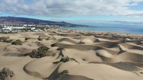 Magnificent aerial view of the sands of Maspalomas Dunes in the island of Gran Canaria, and a calm blue Atlantic ocean under a cloudy sky. Luxury hotels and mountains in the background