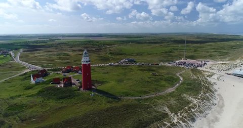 4K Aerial drone shot, slowly flying away from red lighthouse on the white sandy beach of Texel. Birds eye view horizontal wide angle shot bright blue sky with big white clouds, sunny landscape