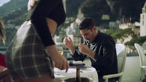 At restaurant with view of coast in the background, a cheating boyfriend sitting with another girl has a drink splashed in his face by his current angry girlfriend. Wide shot on 8k helium RED camera
