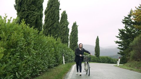 Content Italian man walking along the street with a bike while looking at the trees alongside the road on an overcast day. Wide shot on 8k helium RED camera. วิดีโอสต็อก