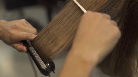 Hairstyling long hair with hair iron in professional beauty studio. Hairstylist doing straightening for long haired woman in barbershop. Hairdresser creating hairstyle brunette woman