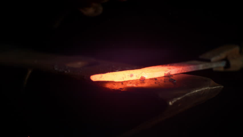 SLOW MOTION, MACRO, DOF: Small black particles flying away from hot red blade while getting forged by an unrecognizable blacksmith. Glowing piece of iron being forged into blade. Forging knife blades. Royalty-Free Stock Footage #1023034696