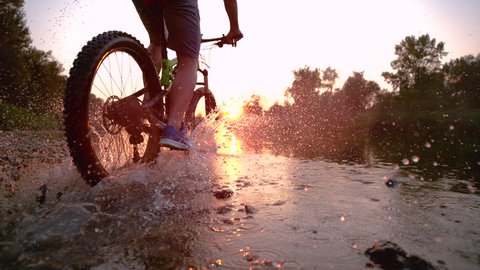 SUPER SLOW MOTION CLOSE UP: Sportsman riding his bicycle in the shallow stream towards the breathtaking golden sunset. Unrecognizable young man riding a mountain bike and splashing cold river water.