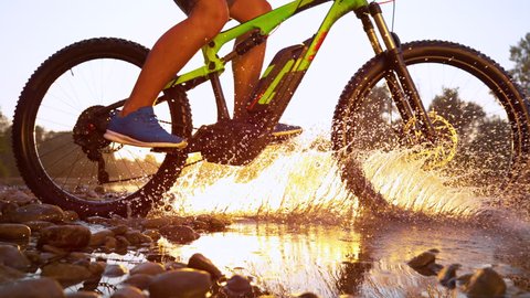 SLOW MOTION, LENS FLARE, CLOSE UP: Unrecognizable cross country cyclist splashes the refreshing river water as he speeds through the picturesque serene nature at sunset. Sportsman riding his bike.