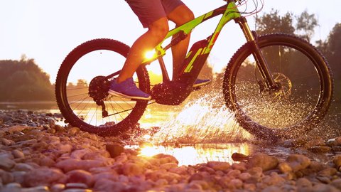 SLOW MOTION TIME REMAP SUN FLARE CLOSE UP: Unrecognizable sportsman riding a mountain bike in a shallow stream running through tranquil nature at sunset. Mountain biker splashing refreshing water.