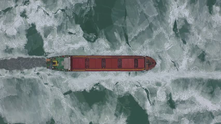 Cargo ship sailing on frozen sea in extreme winter conditions aerial shot. Sailing in narrow fairway channel made by icebreaker vessel. Water transportation during cold winter season in north.