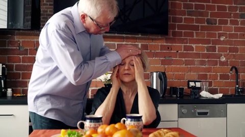 Senior woman having head pain sitting with husband in the kitchen
