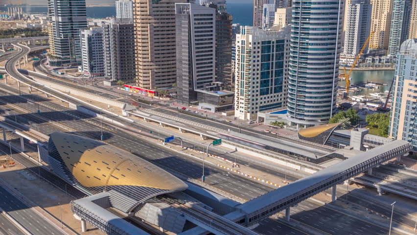 Futuristic building of Dubai metro and tram station and luxury skyscrapers behind timelapse in Dubai Marina, United Arab Emirates. Aerial top view from JLT with traffic on sheikh zayed road