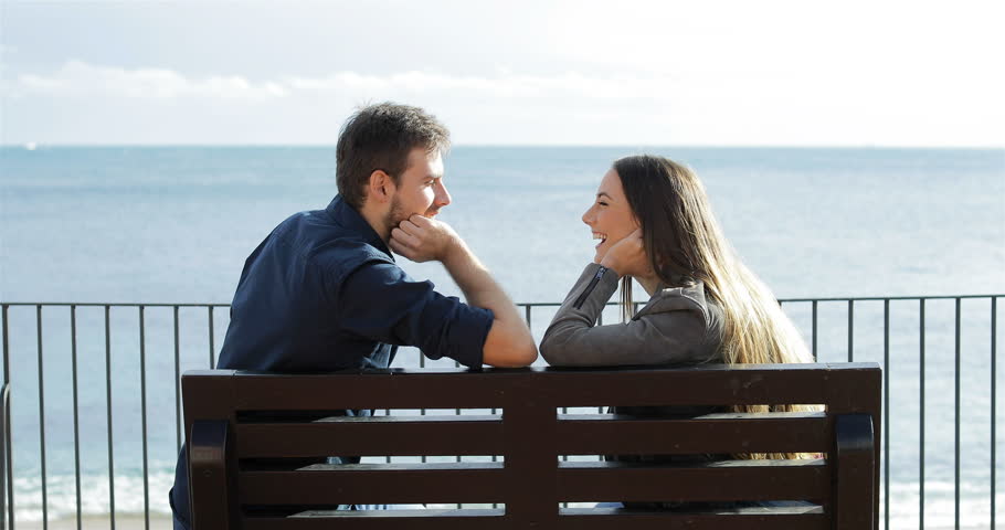 Profile of a happy couple looking each other sitting on a bench on the beach