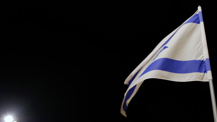 4K. Flag of the State of Israel develops on a flagstaff on wind at night against the background of the black sky and in the left bottom corner of a shot the lamp is visible. | Shutterstock HD Video #1023093352